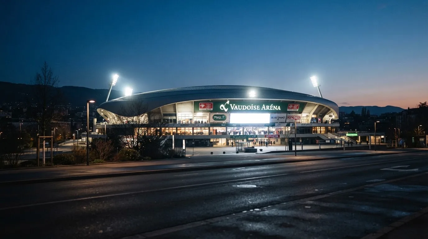 Ein grosses Eisstadion aus der Ferne gesehen, hell erleuchtet von Flutlicht in der Abenddämmerung, davor eine leere Strasse.
