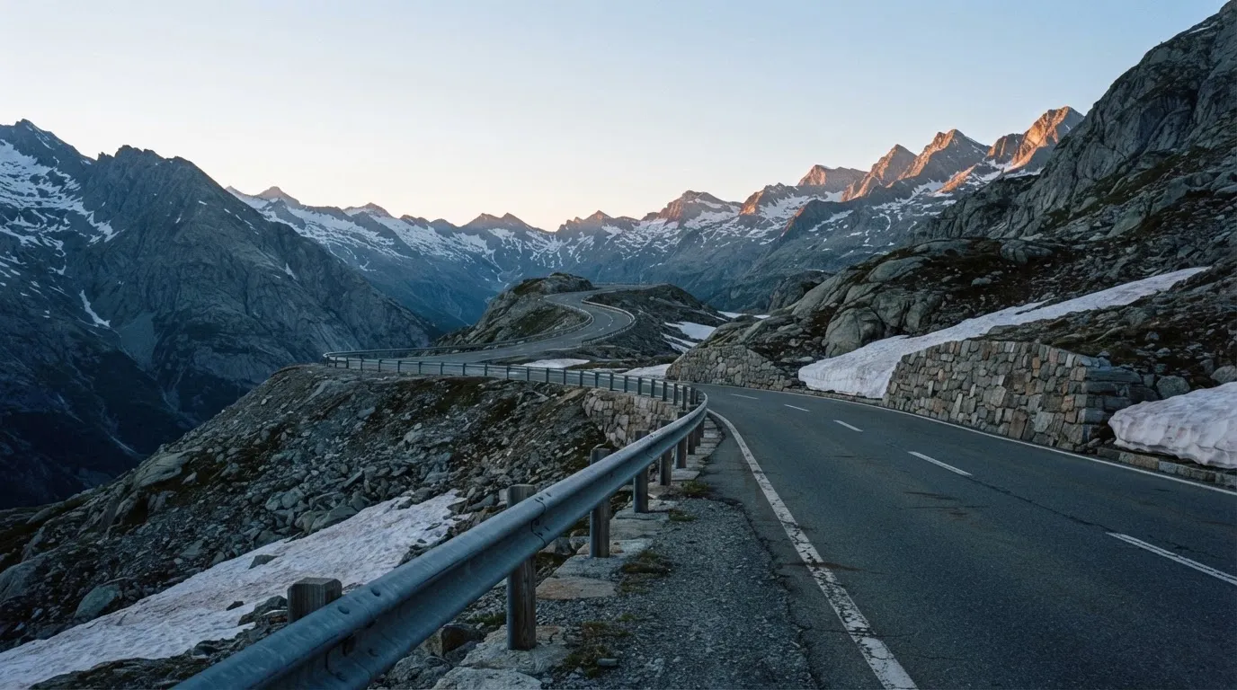 Ein Bergpass in den Schweizer Alpen mit leerer Strasse und Schneeresten am Rand im ersten Frühlicht.
