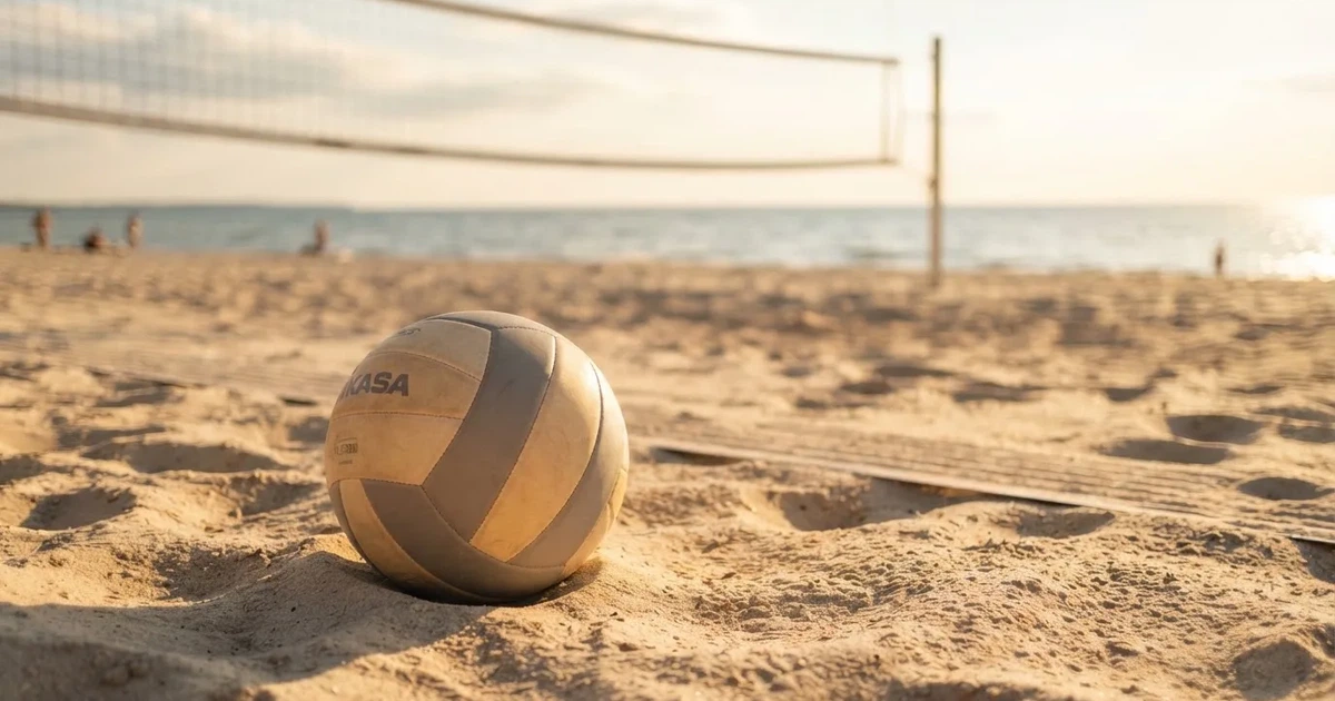 Ein Volleyball liegt auf feinem hellem Sand, ein gespanntes Netz verschwimmt dahinter, das Licht kommt von einem weiten Meer.