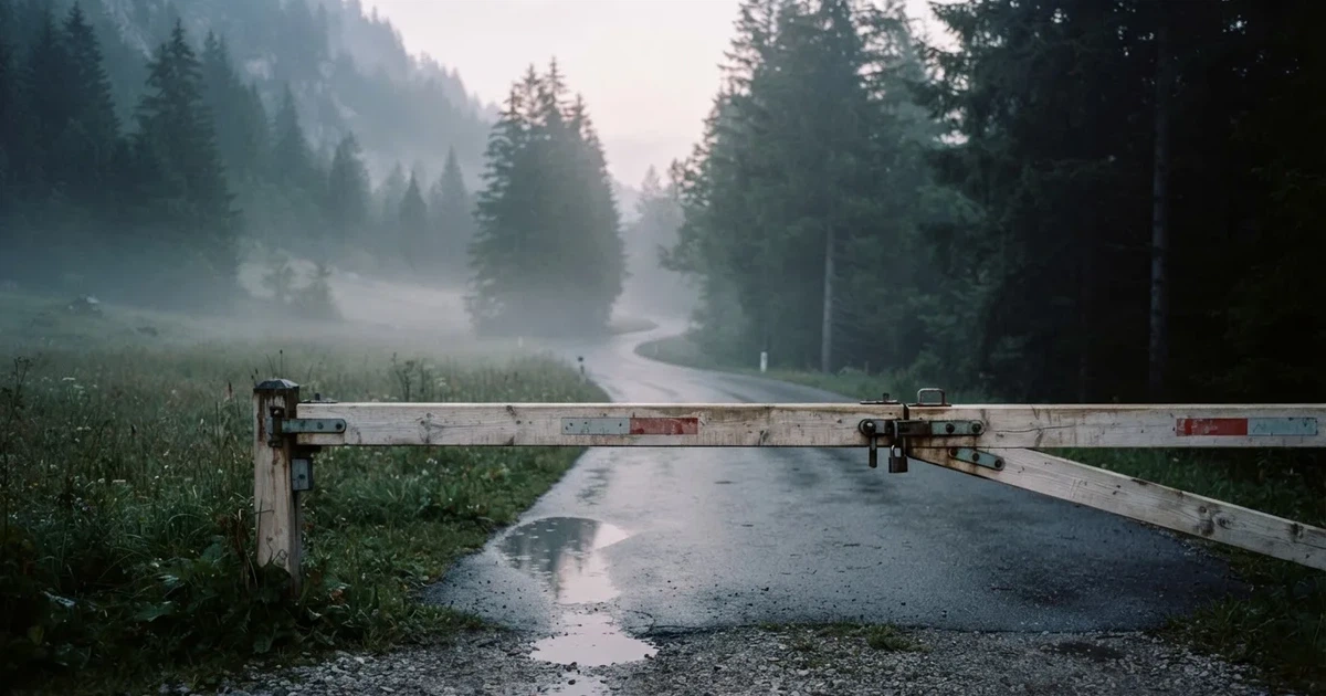 Eine geschlossene Schranke steht an einer ruhigen Bergstrasse im Morgennebel, dahinter Tannen und eine nasse Wiese.