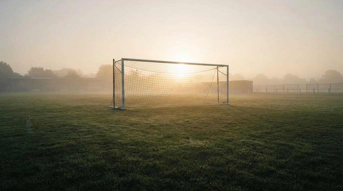Ein Torpfosten mit Netz steht im Gegenlicht auf einem leeren Fussballplatz, über dem Rasen liegt Nebel.